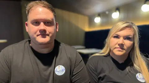 Darren, a man with short light brown hair, wearing a black T-shirt. He is sitting next to Jemma, a woman with long blonde hair, wearing a black T-shirt. Neither are smiling. 