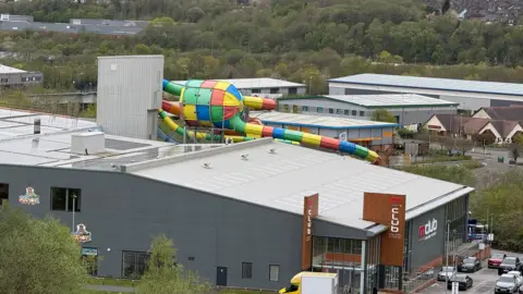 A large building with branding indicating it is a gym. There is a multi-coloured water slide at the back of the building and further back are other industrial buildings and a pub. There are also a number of trees in the distance.
