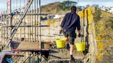 A man carries two yellow buckets of mortar up some stone steps. He is wearing a dark blue jumper. There is scaffolding to his left. 