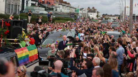 PA Media A huddle of people stand on the street as a hearse covered in flowers drives through the street