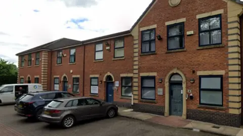 Google The large red-brick building has black window frames and doors and a yellow brick trim around them. There are several cars parked outside.