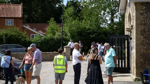 Reuters. Residentes evacuados se congregan en el ayuntamiento de Westleton. Es un día soleado. Una persona lleva un perro. Varios grupos conversan fuera del ayuntamiento.