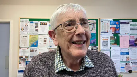 An elderly man with short white hair and glasses is wearing a grey sweater over a check shirt. He is standing in front of a noticeboard.