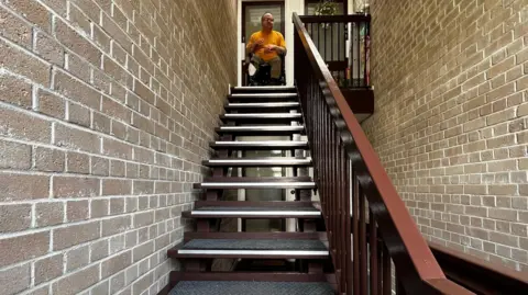 James Smith, a man in a wheelchair, is sitting in the open front door of his flat at the top of a flight of stairs. The stairs are open stairs - so there is space between each one - and a wooden handrail is to the right. 