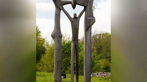 Nene Park Trust The Peterborough Arch is an abstract sculpture of a wooden man standing under a wooden arch.
