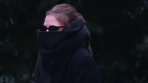 Ellis Eyles walking outside of court. She is wearing large black sunglasses and a black scarf that covers most of her face. She has brown hair which is tied back and is wearing dark clothing.