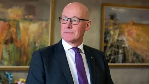 John Swinney wearing a dark suit over a white shirt with a purple tie. He is standing in front of two paintings on a neutral background.