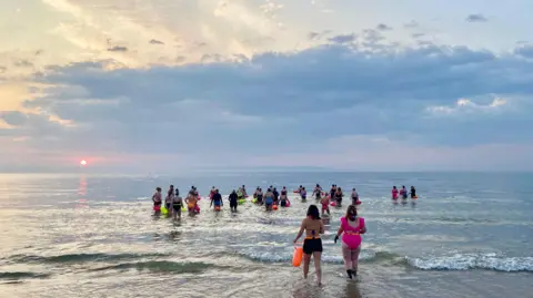 Elena Reid A group of swimmers, wearing multi-coloured swimwear walking through the blue and waves waves into a sea. The sky above is cloudy and blue, in the distance the a pink-toned sun is setting.