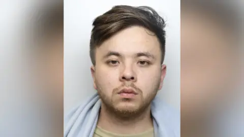 National Crime Agency Joshua Ee's mugshot. He has brown hair which is long on the top and short on the sides and stubble around his chin. He is looking to the left of the camera with a straight face and looks to have a blanket around his shoulders.