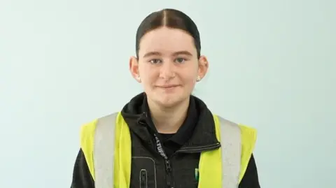 BBC/Qays Najim A teenage girl with dark hair scraped back in a ponytail stands in an empty room with light blue walls. She is wearing a dark jacket with a hi-vis vest on top and holds a spanner in her hands