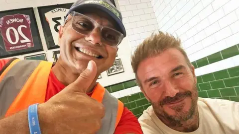 Pesh Kapashiawala is wearing an orange hi-vis vest over a red T-shirt. He is smiling and giving a thumbs up next to Sir David Beckham, who is wearing a white T-shirt and sitting down inside the restaurant. Sir David is also smiling.