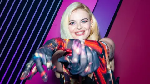 A woman with blonde hair wearing a multicoloured dress points at the camera while smiling in front of a pink neon backdrop.