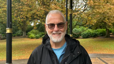 BBC Campaigner smiles at the bandstand in Cannon Hill Park. He is wearing a blue top and black jacket. 