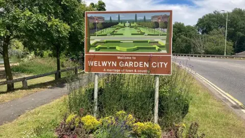 Brian Farmer/BBC A brown sign showing a picture of a formal garden above the words "Welcome to Welwyn Garden City, a marriage of town and country" in white letters. It is set in a bed of shrubs surrounded by grass with a tree-lined path on one side and a road with double yellow lines on the other.