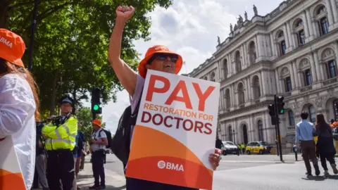 Getty Images A junior doctor holds a placard that says "Pay restoration for doctors" during a rally in London in June last year. They are holding up one arm and wearing an orange British Medical Association hat and sunglasses, while their body is largely obscured by their sign. Behind them other protesters, a police officer and pedestrians are standing around.