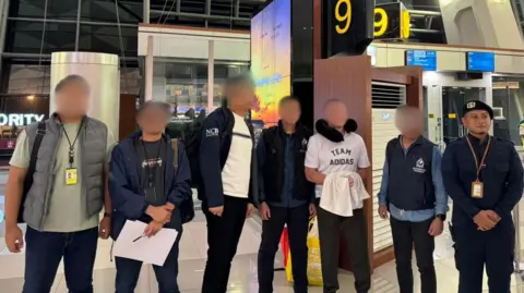 Ngurah Rai Immigration Office A line-up of seven men in an airport. They wear casual clothes and all of them have their faces blurred out except an official on the far right. Steven Lyons is third from the left and wears a white t-short which reads "Team Adidas", black trousers and a black neck pillow. He holds a white item of clothing 