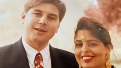 Kam Miller and Neil Miller. Neil is wearing a shirt and tie and has short hair. While Kam is wearing jewellery and has long brown hair. 