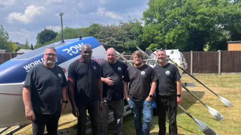 Frank Bruno stands with the group that are rowing the Atlantic, with the boat behind them and oars to one side. Left to right, the men seen in the picture are Rob Oliver, Matt Humphries, Tom Hayward and Kev Joynes.
