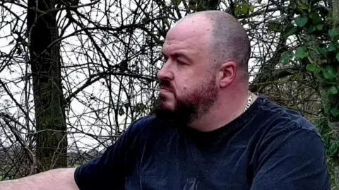 A man with a dark beard and shaved head in a black tshirt in a woodland area