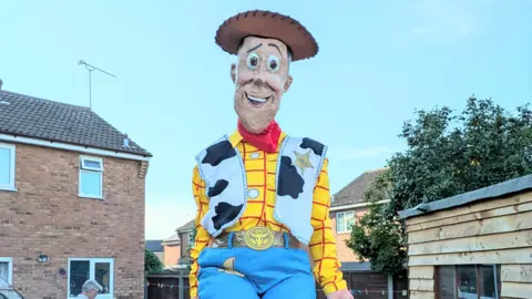 Contributed A model of Woody the cowboy from the animation, Toy Story. The model is wearing a brown cowboy hat, yellow and red chequered shirt, a jacket with a cow print on it and blue trousers. The model is sitting atop a garden fence. 