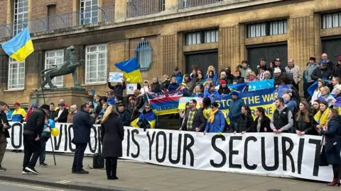 Aimee Dexter/BBC People are standing on the steps which lead up to City Hall in Norwich. Many are holding Ukrainian flags. A speaker holding a microphone is standing in front of them.