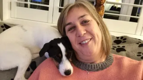 Donna, who has short brown and grey hair, smiles at the camera. She is wearing a pink jumper with a grey neckline and is sitting on the sofa with a small white and brown dog, who is curled up against her. 