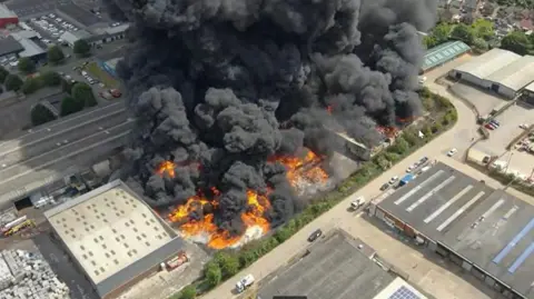Northamptonshire Fire and Rescue Service An aerial shot of a large recycling warehouse fire, showing flames and large amounts of smoke, buildings around it and cars on the road.
