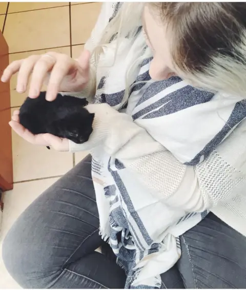 A woman holds a small black kitten in her hand as she strokes it 