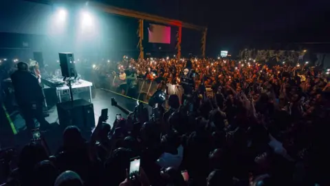 A huge crowd of people inside a music venue, some holding their phones aloft, watching a DJ at their decks, with barriers separating them from the crowd. Speakers can be seen either side of the decks.