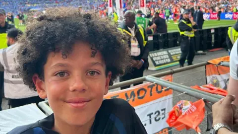 A boy is seen smiling at the camera. He is wearing a football jersey and there are people in the background at a football match