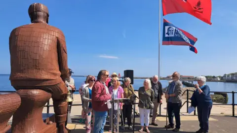 Brian Chambers A group of people stand on the pier looking on at one person speaking on a microphone. The back of a Fiddler's Green Fishermen's Memorial is on the left, which is a tall brown sculpture depicting a man wearing a flat cap sitting and looking out to sea. Two flags fly behind the group. On red flag with a union jack in the corner and another red and blue one which reads: "The Fishermen's Mission."