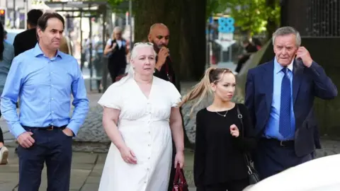 PA Media Neville Leeson, wearing a light blue dress shirt and dress pants walks along on a street next to a woman wearing a white dress, another woman wearing a black jumper and an older man wearing a blue dress shirt and navy shirt and tie. 