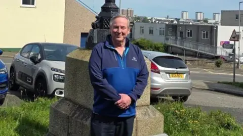 Local Democracy Reporting Service A close up of Rob Parsonage. He has short grey hair and is wearing a blue fleece. He is standing in front of the memorial on which the lantern sits.