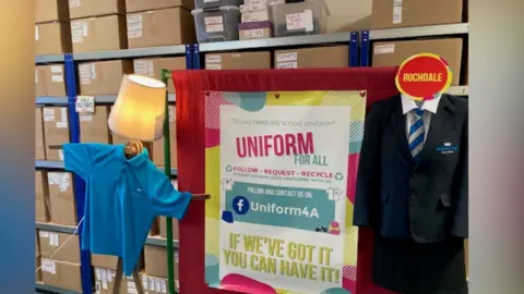 A sign 'Uniform for All' and two uniforms hung up next to it with many cardboard boxes stacked on shelves in the background.