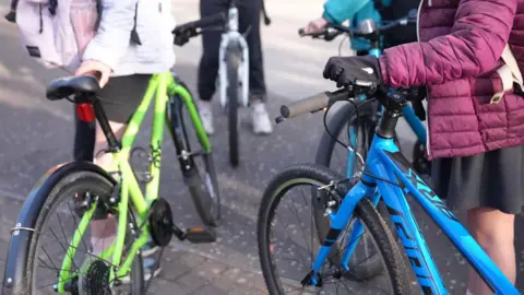 Two children stand next to bicycles. One bike is green and the other is blue. One of the children has a purple coat on and the other has a white coat on.
