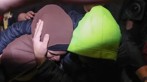 A hijabi woman is shielded from media by a man wearing a bright green hoodie