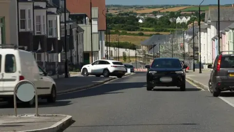 Several cars in the foreground of a wide shot of a road through Nansledan. There is a white road bridge, and countryside fields and trees with a blue sky in the background. The road has houses being built with scaffolding on the right hand side  and shops on the left.