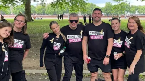 North West Anglia NHS Foundation Trust Seven members of Abi Quinlisk's family. They are all wearing black T-shirts saying Young Lives vs Cancer. Behind them is a park, showing grass and trees.