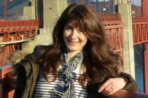 Dana Dawson Professor Dana Dawson smiling at the camera, she has long brown hair and is wearing a white top with blue stripes and a green jacket, there is a bridge in the background.