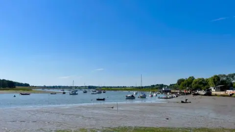 Martin Giles/BBC A picture of the River Deben from the quayside in Woodbridge.  The sky is big and blue the tide is out so there some mud flats and then the river. On the rivers there are some boats bobbing in the distance. 