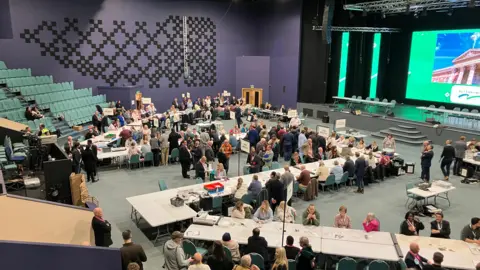 A widescreen shot, taken from a balcony, of the election count, with many people sat at tables and party members chatting. There is a stage on the right.