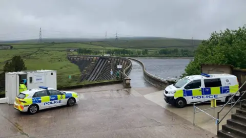 Police vehicles in the foreground, with a dam and reservoir in the background.
