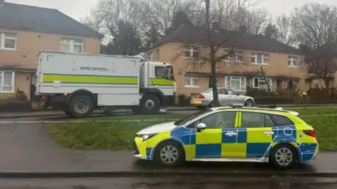 A police car is parked outside a row of beige houses next to a green. The sky is grey and the street is wet.