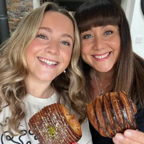 Jessica Morgan-Helliwell Jessica (on the left) and her mother Louise (on the right) are smiling in a selfie. They're holding a croissant each up to the camera. Jessica has blonde hair and is wearing a white t-shirt and Louise has brown hair and is wearing a black top.