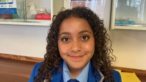 Connie. She has long dark brown curly hair, brown eyes and wearing a blue school uniform.