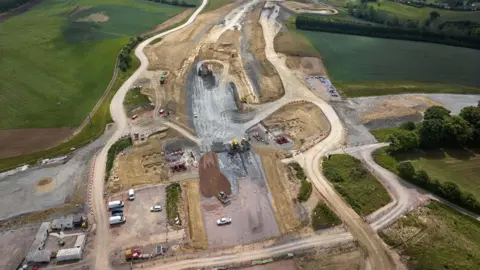 An aerial picture shows a major road construction site in the middle of green fields. A huge amount of groundwork has been undertaken, with a number of dirt roads encircling the site. Work vehicles, including white vans and yellow trucks, appear tiny.