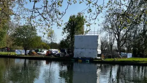 The River Great Ouse, with a bank on the other side of the river, with lots of trees, a guillotine block all covered in white covering and scaffolding, also machinery and vehicles around the building. 