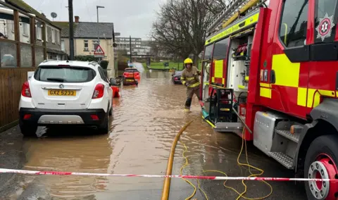 A picture of a flooded street - on the right is a fire engine, with a hose coming out of its rear. A firefighter stands at the back of the truck. On the ground is a foot or so of dirty brown water. On the left are parked cars and a row of terraced housing. 