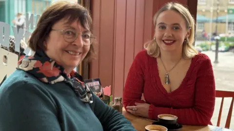 Boston Tea Party A close up of Barbara, wearing a teal coloured jumper and a floral scarf and her daughter, wearing a red frilly top and a striking silver pendant necklace, sitting on a coffee shop table with cappuccinos in front of them and windows onto the street behind. Both are smiling to camera.