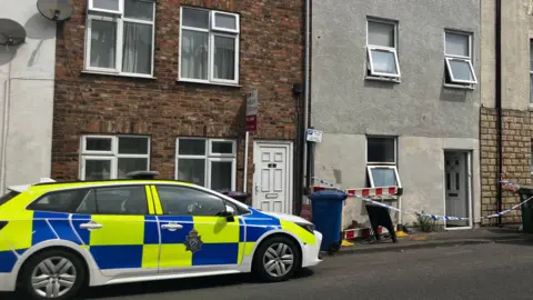 A bright yellow and blue police car with a star logo is parked next to a grey terrace house which has a white and blue striped police cordon draped across it. All the doors and windows of the property are open.